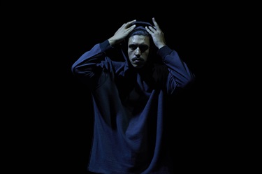 A tall Aboriginal man wears a dark hooded jumper and raises his hands to his head to peel off the hood.